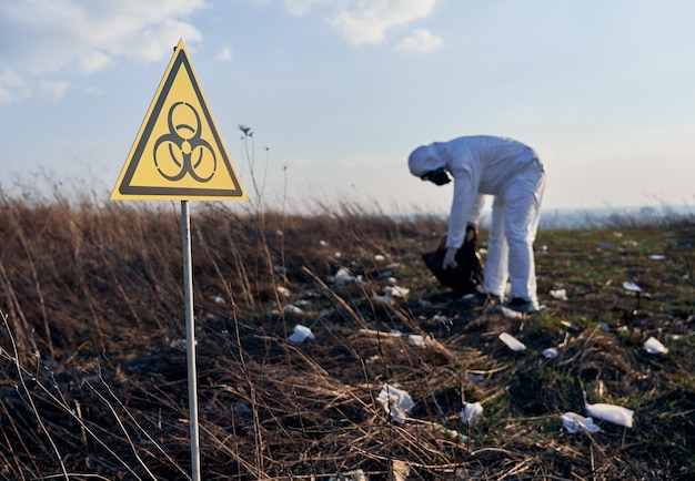 Foto gratuita ricercatore in tuta protettiva che raccoglie immondizia di plastica nel sacco nero dei rifiuti all'aperto in una giornata di sole