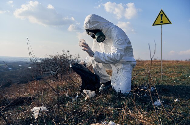 Researcher in protective suit collecting plastic garbage into black waste bag outdoors on sunny day