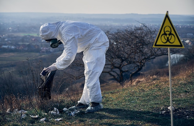 Foto gratuita ricercatore in tuta protettiva che raccoglie immondizia di plastica nel sacco nero dei rifiuti all'aperto in una giornata di sole