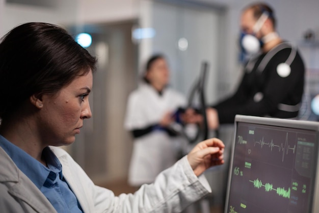 Researcher woman analyzing heart rate pulse on monitor while in background runner man working at body endurance running on fitness trainer in modern laboratory. Specialist doctor monitoring EGK data