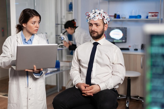 Researcher woman doctor typing neurological disease symptoms on laptop discussing medical treatment with patient. Neurologist engineer monitoring nervous system activity of man with eeg headset