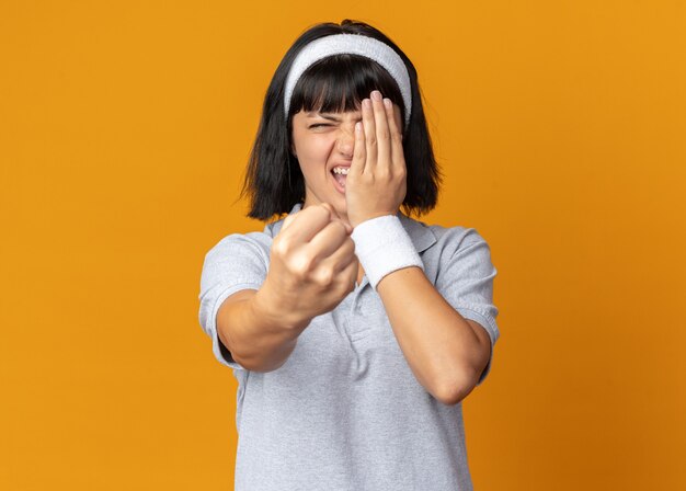 Resentful and angry young fitness girl wearing headband showing fist covering her eye with hand 