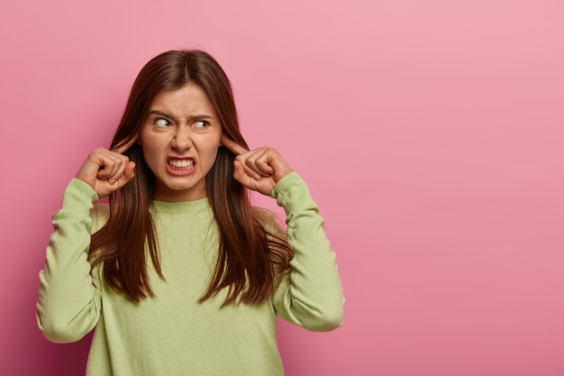 Foto gratuita la donna infastidita risentita tappi le orecchie, irritata dal rumore forte, evita il suono fastidioso, vestita con un maglione verde casual, stringe i denti, aggrotta le sopracciglia, isolato sul muro rosa, spazio vuoto a parte