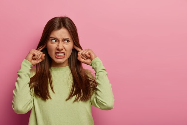 Resentful annoyed woman plugs ears, irritated with loud noise, avoids annoying sound, dressed in casual green jumper, clenches teeth, frowns face, isolated on pink wall, blank space aside