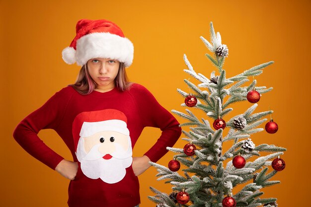 Resentful little girl in christmas sweater and santa hat looking at camera with angry frowning face with arms at hip standing next to a christmas tree over orange background