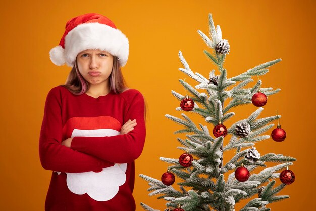 Resentful little girl in christmas sweater and santa hat looking at camera with frowning face with arms crossed standing next to a christmas tree over orange background