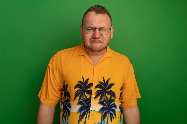 Free photo resentful man in glasses and hawaiian shirt with sad expression pursing lips standing over green wall