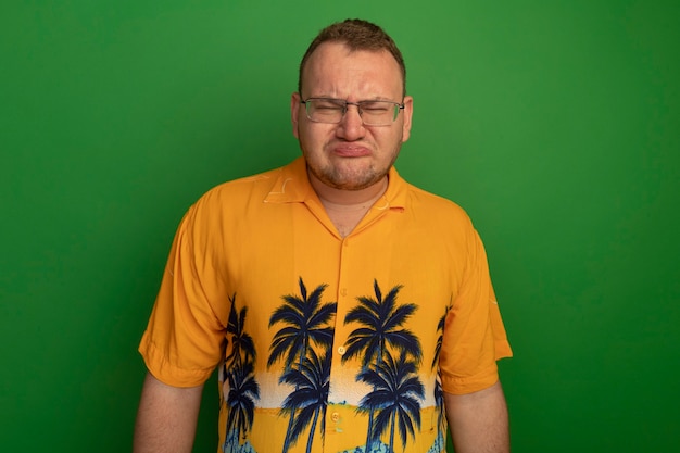 Resentful man in glasses and hawaiian shirt with sad expression pursing lips standing over green wall