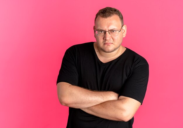 Free photo resentful overweight man in glasses wearing black t-shirt with frowning face with crossed arms over pink
