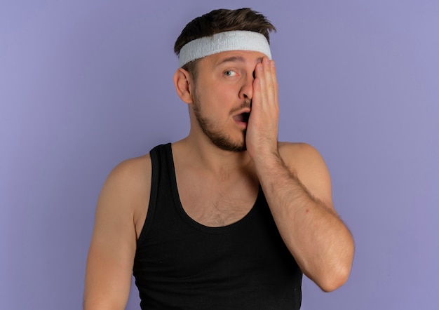 Free photo resentful young fitness man with headband covering one eye wirh arm standing over purple background