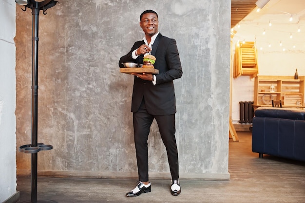 Respectable young african american man in black suit hold tray with double burger against gray wall