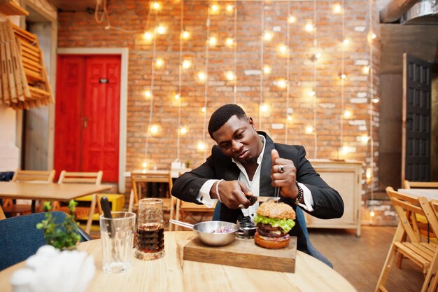 Respectable young african american man in black suit sitting in restaurant with tasty double burger and soda drink Divide half by knife