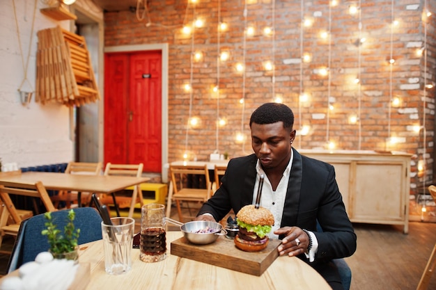 Respectable young african american man in black suit sitting in restaurant with tasty double burger and soda drink