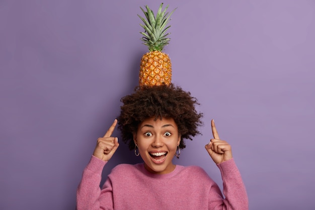 Rest and chill. Look how I can! Joyful curly haired ethnic girl points at ripe pineapple on head, laughs positively