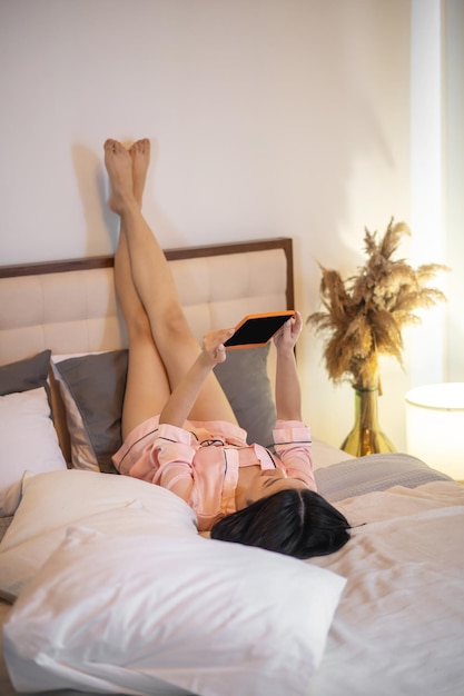 쉬다. 분홍색 잠옷을 입은 흑갈색 머리의 젊은 여성이 밝은 방에 있는 침대에서 화면을 보고 있는 태블릿을 들고 다리를 들고 등을 대고 누워 있습니다.