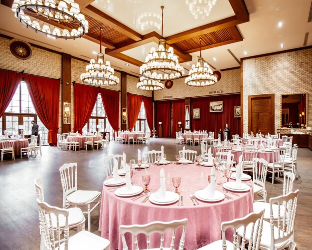 丸テーブル、白いナポレオンの椅子、赤いカーテン、レンガの壁、シャンデリアのあるレストランホール