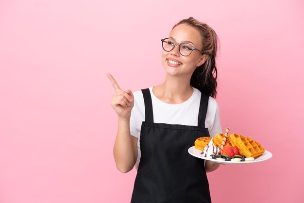 Русская девушка официант ресторана держит вафли, изолированные на розовом фоне, намереваясь реализовать решение, подняв палец вверх