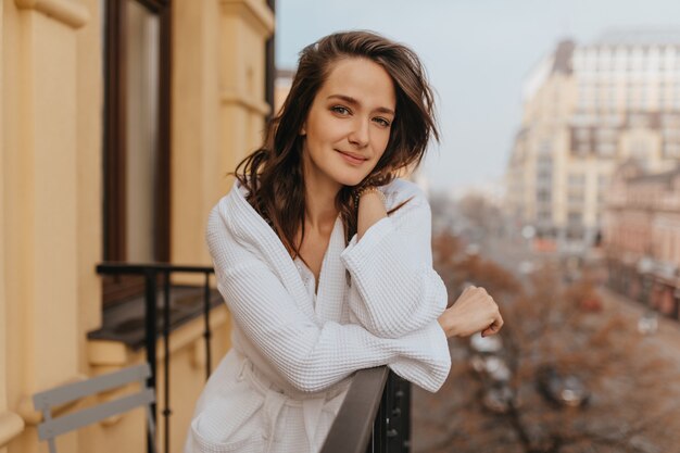 Rested woman without make-up in bathrobe looks into camera, posing on open balcony.
