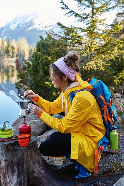 安らかな女性旅行者がキャンプストーブでコーヒーを作り、切り株の近くでポーズをとり、さまよった後に休憩します