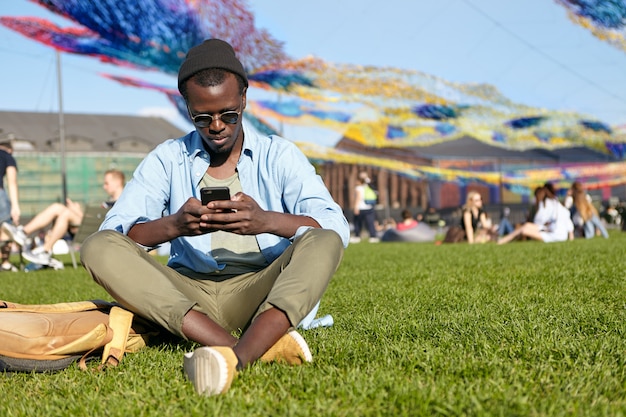 Restful male with dark skin wearing black hat, sunglasses, shirt, trousers and sport shoes, keeping legs crossed, relaxing at green lawn, looking attentively in his mobile phone reading news online