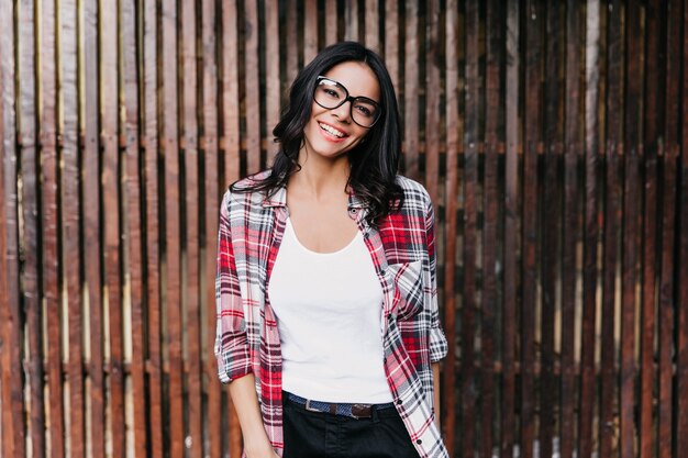 Resting latin woman in glasses standing on wooden wall. Outdoor shot of spectacular girl enjoying spring photoshoot.