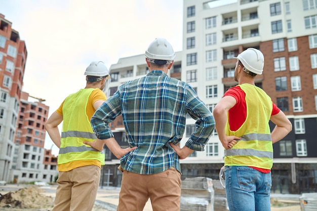 Result. Back view of three men in protective helmets holding hands on his waist standing at construction site looking at buildings under construction during day