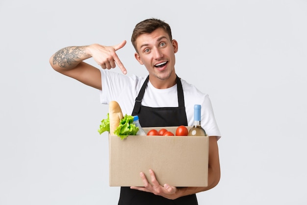 Retail grocery shopping and delivery concept handsome courier in black apron handing over box with g...