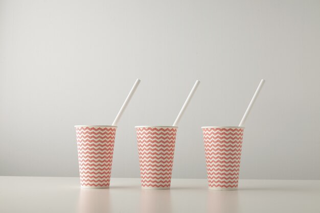 Retail set of three paper cups decorated with red line pattern and with white drinking straw inside isolated on white table