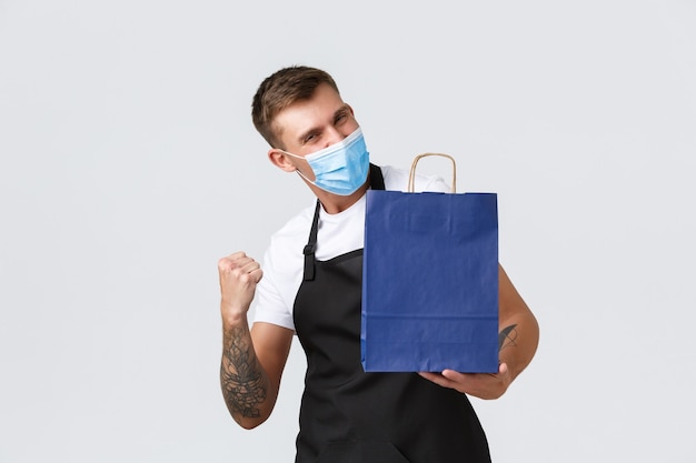 Retail store, shopping during covid-19 and social distancing concept. Enthusiatic handsome salesman working during coronavirus pandemic in shop, fist pump as holding eco bag with purchase