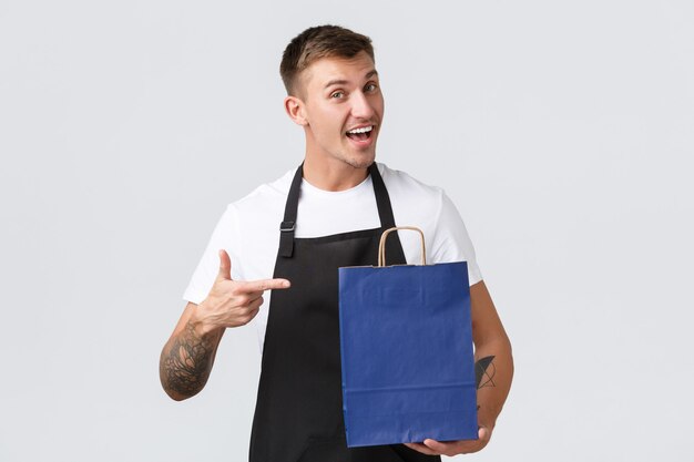 Retail store, shopping and employees concept. Enthusiastic friendly salesman in black apron, pointing finger at eco-bag with clients purchased products, standing white background