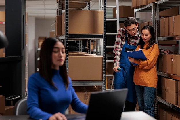 Foto gratuita dipendenti del magazzino al dettaglio che controllano gli appunti con l'elenco degli ordini nel magazzino. supervisore del magazzino che spiega il sistema di inventario delle merci al gestore dei pacchi asiatico con le stampelle