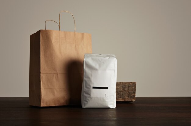 Пакет товаров для розничной торговли: большой герметичный пакет белого цвета с пустой этикеткой рядом с бумажным пакетом и деревенским деревянным кирпичом на красном столе.