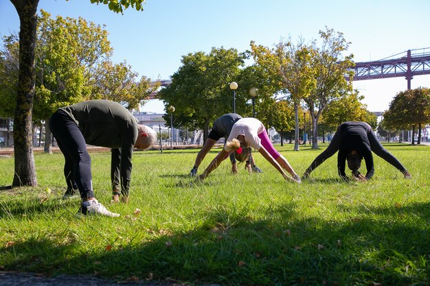 Retired active mature people wearing sports clothes, doing morning exercise on park grass, stretching back and legs muscles. Retirement or active lifestyle concept