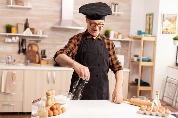 Retired chef in home kitchen wearing apron while sprinkling ingredient on table for tasty