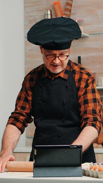 タブレットで料理のアドバイスを受け、ソーシャルメディアで料理のチュートリアルを学び、木製の麺棒で生地を作る引退した男性。ホームケーキを準備するラップトップを使用して骨とエプロンを持つ祖父