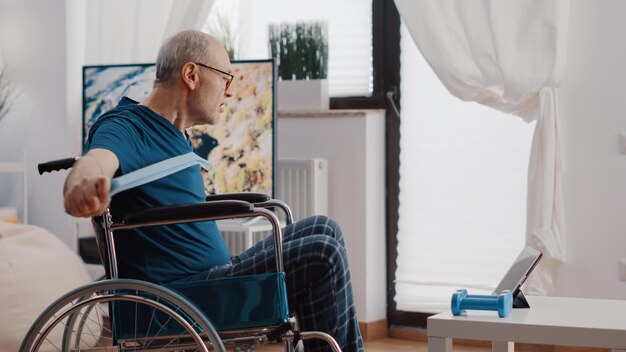Пенсионер сидит в инвалидной коляске и использует эспандер для упражнений, следя за видео тренировки на планшете. Пожилой взрослый с ограниченными физическими возможностями тянет эластичный ремень, чтобы растянуться