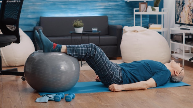 Retired person using fitness toning ball and lifting legs to stretch muscles. Aged man training and doing physical exercise while laying on yoga mat. Pensioner exercising with equipment