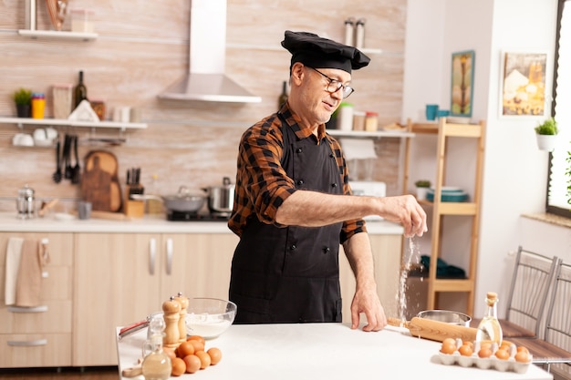 Retired senior baker wearing apron and bonete using ingredients for homemade pizza