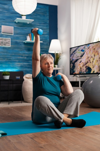 Retired senior woman sitting on yoga mat in lotus position raising hand during wellness routine warming up training body muscles using dumbbells
