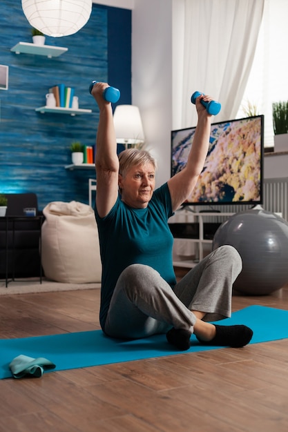 Retired senior woman sitting on yoga mat in lotus position raising hand during wellness routine