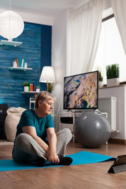 Retired senior woman in sportswear sitting on yoga mat watching fitness lesson on laptop