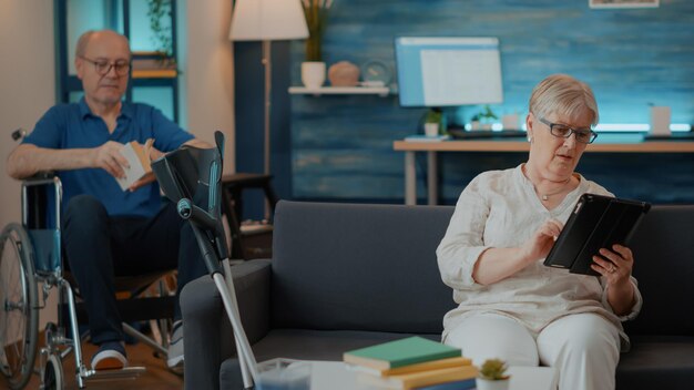 Retired woman browsing internet on digital tablet at home. Elder person with crutches on sofa, using modern gadget with touch screen to learn technology in living room. Pensioner with device