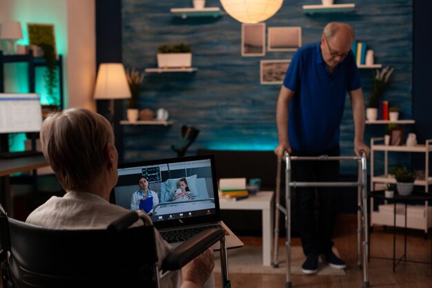 Retired woman holding laptop with video call conference talking to doctor from hospital and niece. Old adult sitting in wheelchair while senior man using walk frame in living room background