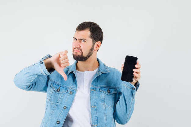 Retro-style man in jacket,t-shirt showing thumb down while showing phone and looking displeased , front view.