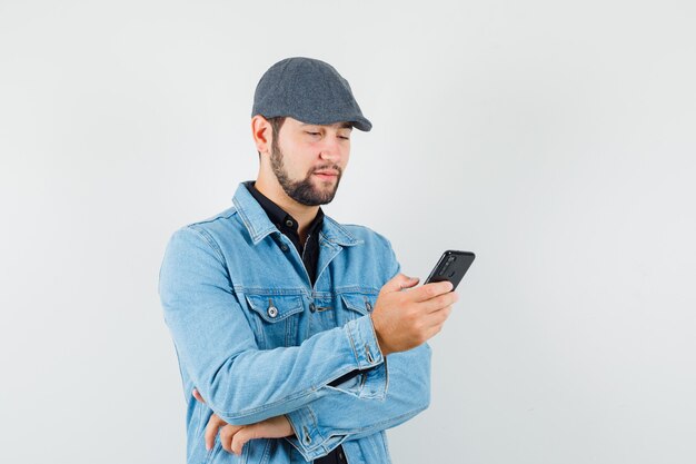 Retro-style man looking at phone in jacket,cap,shirt and looking concentrated. front view.