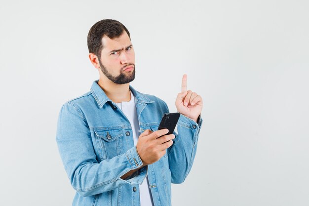 Retro-style man pointing away while holding his phone in jacket,t-shirt and looking displeased , front view. space for text