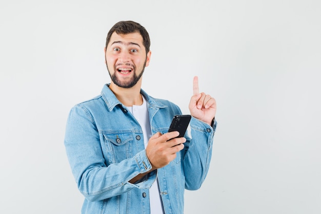 Retro-style man showing new idea gesture while holding his phone in jacket,t-shirt and looking hopeful , front view.