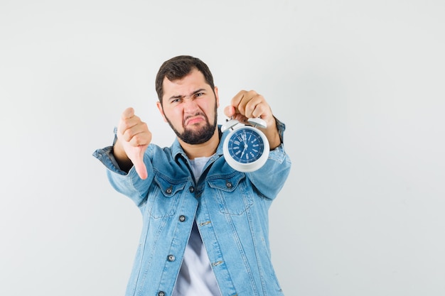 Retro-style man showing thumb down while holding clock in jacket,t-shirt and looking dissatisfied , front view.
