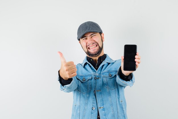 Человек в стиле ретро показывает палец вверх, показывая телефон в куртке, кепке, рубашке и выглядит счастливым, вид спереди.