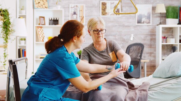 Revealing shot of female nurse helping an elderly woman to recover her muscles after an accident. She is lying in a hospital bed in a retirement home